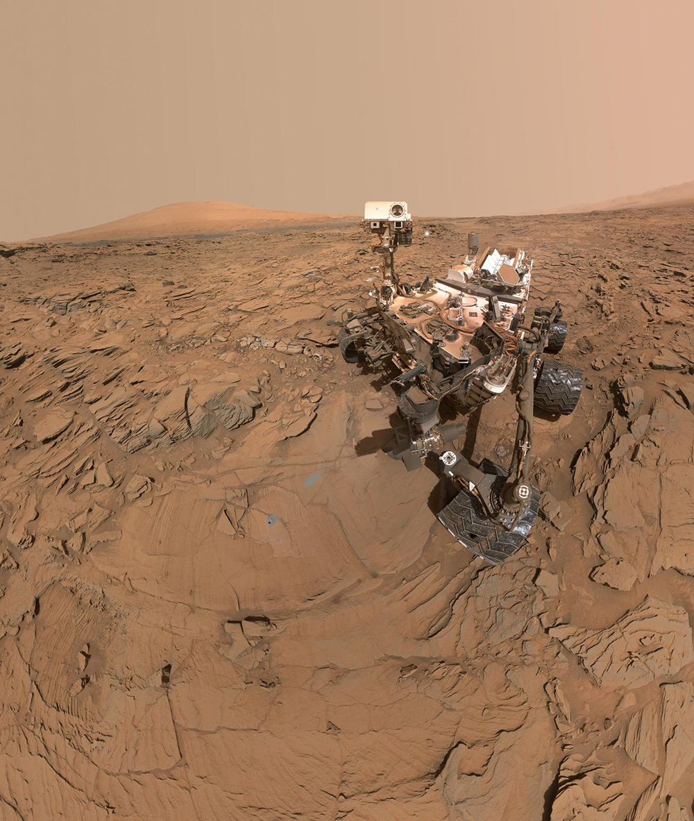 The Mars Curiosity Rover faces the camera, surrounded by red, rocky Mars terrain