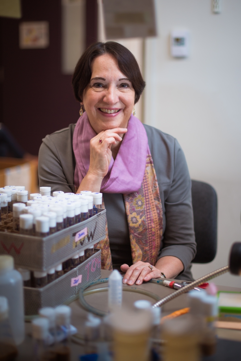 A woman in a pink scarf smiles at a lab bench.