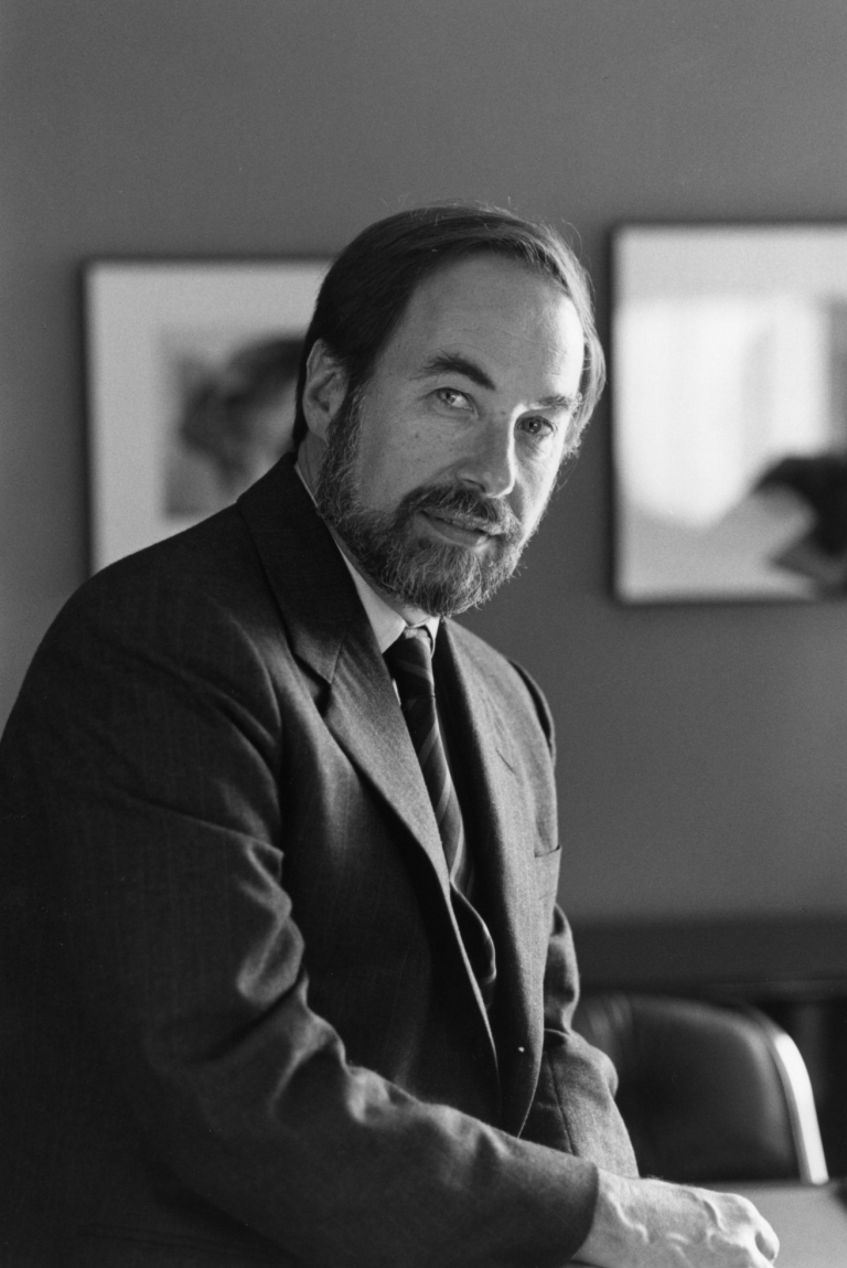 A bearded man in a suit sits with his hands on his lap.