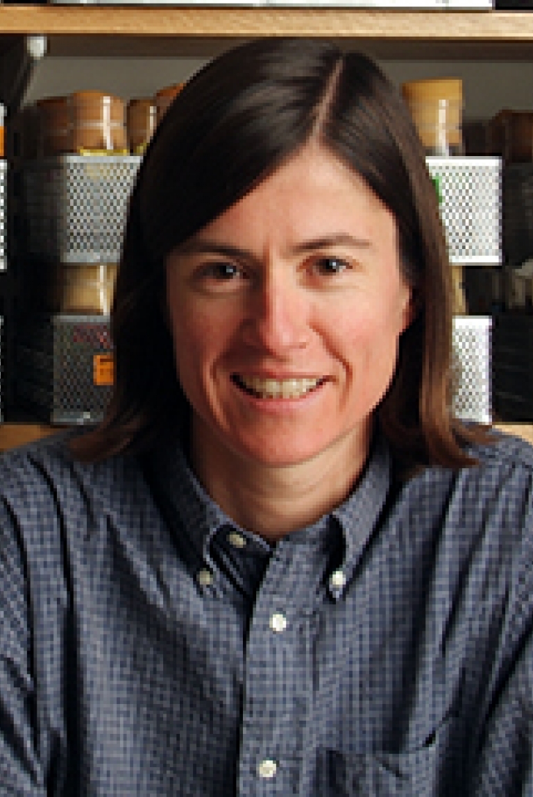 A woman in a blue button-down smiles in front of a wall of bottles.