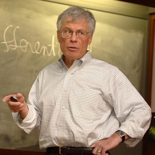 A man in a button-down stands at a blackboard with a piece of chalk. 