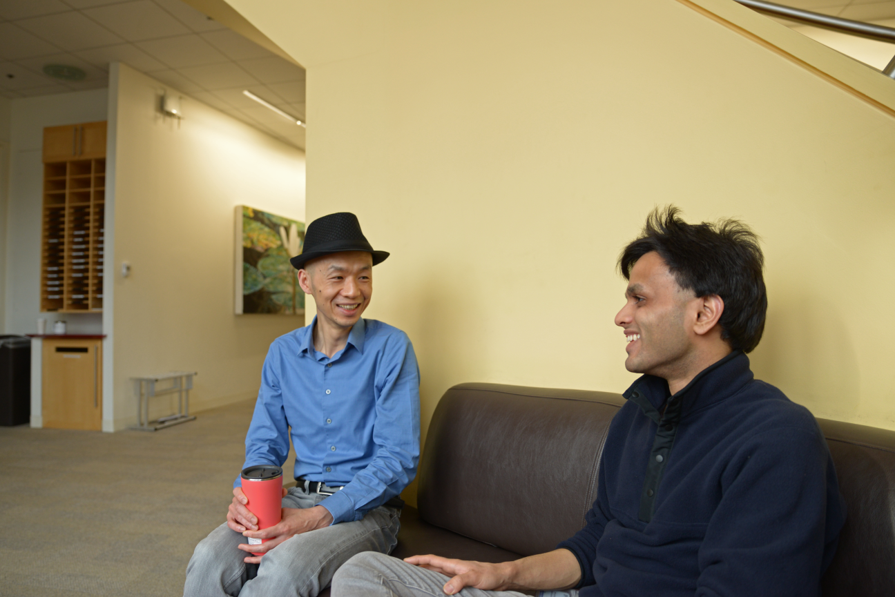 Two men sit turned towards each other on a couch, smiling.