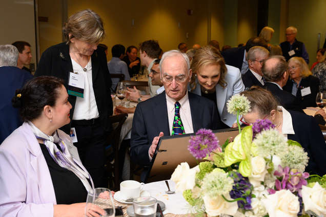 Gerry Fink sitting at a table with a center bouquet and looking at a large plaque that he's holding while Ruth Lehmann and others look on beside him