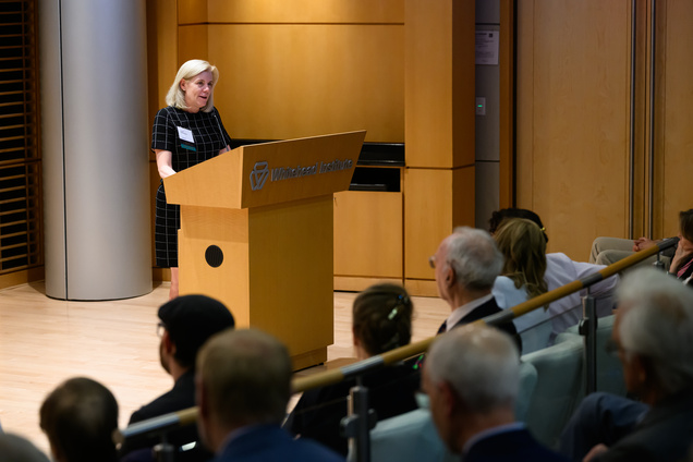 A woman with the nametag "Sarah Williamson" speaking at the podium while Fink and other audience members watch