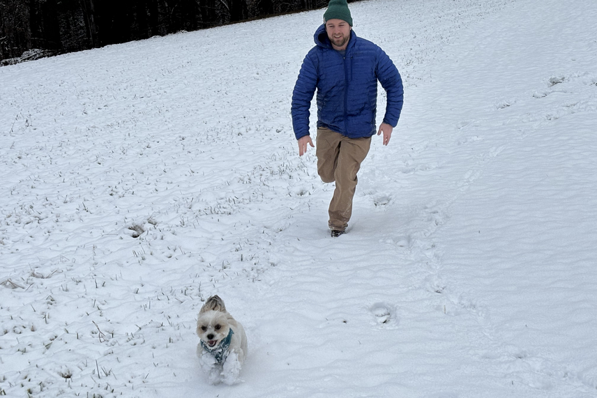 A small, fluffy white dog running through a snow-covered field towards the camera, with Henry running after the dog